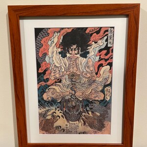 May include: Framed Japanese woodblock print featuring a seated figure with a fierce expression, surrounded by flames and mythical creatures. The artwork is in shades of red, black, and cream, and is presented in a dark wood frame.