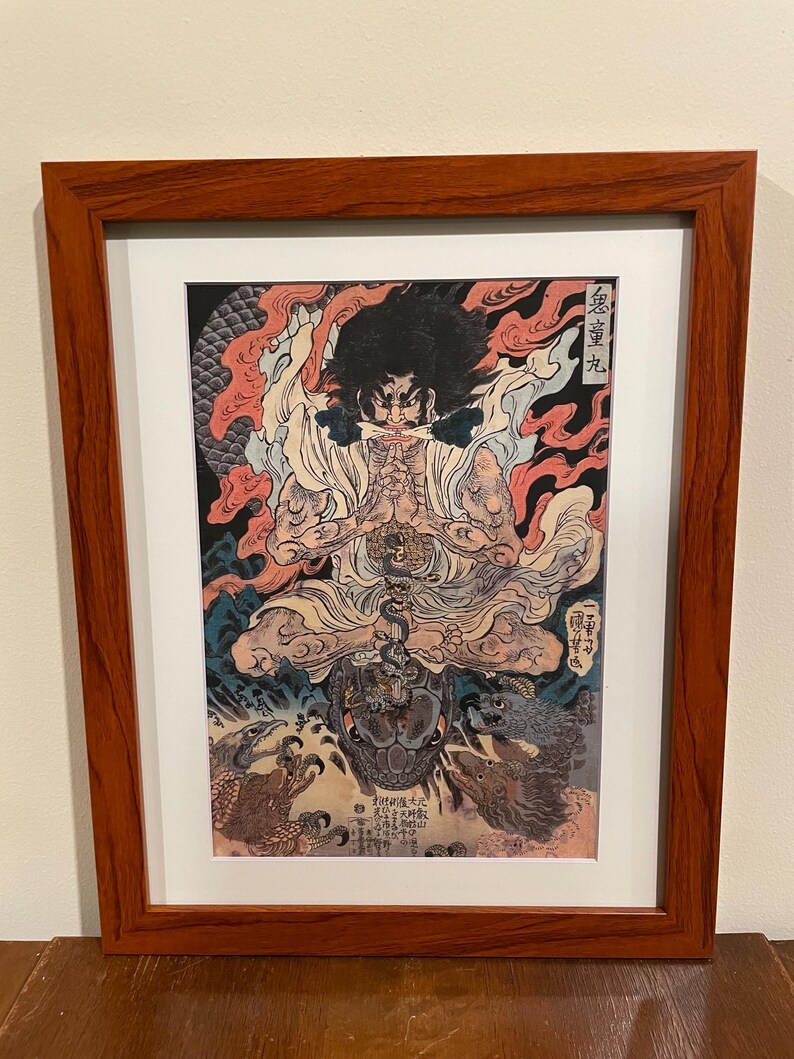 May include: Framed Japanese woodblock print featuring a seated figure with a fierce expression, surrounded by flames and mythical creatures. The artwork is in shades of red, black, and cream, and is presented in a dark wood frame.