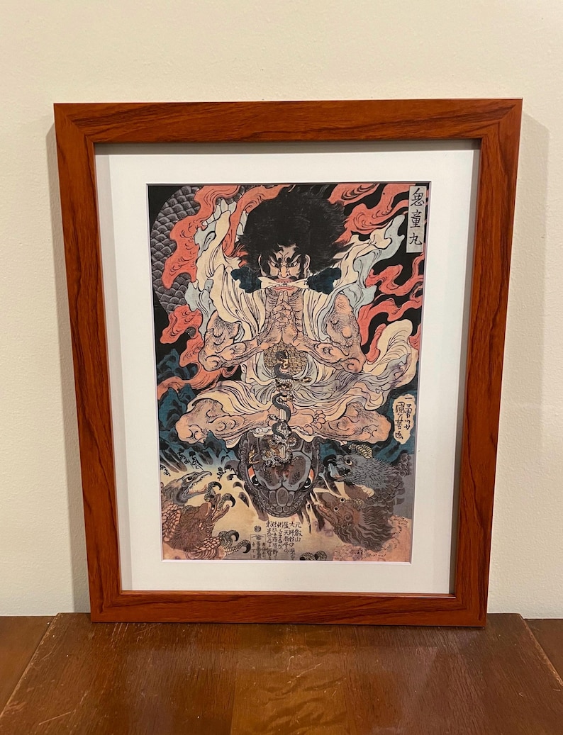 May include: Framed Japanese woodblock print featuring a seated figure with a fierce expression, surrounded by flames and mythical creatures. The artwork is in a wooden frame with a white mat.