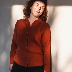 Könnte beinhalten: Eine Frau mit lockigem braunen Haar lächelt und trägt ein rostfarbenes Hemd mit Knöpfen und eine schwarze Hose.