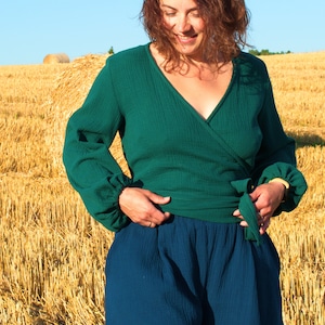 May include: A woman wearing a green wrap top with long sleeves and a blue pair of shorts. She is standing in a field of hay.