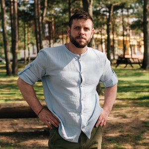 May include: A man wearing a light blue button-down shirt with a mandarin collar. He is standing in a forest with trees in the background.