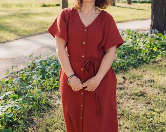 Puede incluir: Una mujer lleva un vestido midi de color óxido con botones en la parte delantera y un cinturón de lazo. El vestido tiene mangas cortas y cuello en V.