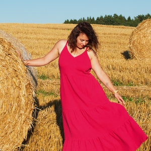 May include: A woman in a bright pink maxi dress stands in a field of hay bales. She is looking down at the ground and her hair is long and brown.