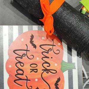 May include: A black and white striped pumpkin with the text "Trick or Treat" in black script. There are black bats flying around the pumpkin. The pumpkin is on a wooden sign with a black and white striped background.