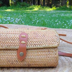May include: A woven straw crossbody bag with a brown leather strap and a brass buckle closure. The bag is sitting on a wooden surface.
