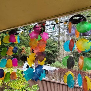May include: A colorful wind chime made of painted, round, and leaf-shaped pieces of metal hangs from a porch roof. The wind chime is made up of multiple strands of metal pieces, each strand with a different color scheme. The wind chime is hanging in front of a window with a view of trees.