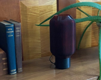 May include: A dark red cylindrical lamp with a ribbed texture and a black base. The lamp is sitting on a wooden surface next to a potted plant with green leaves.