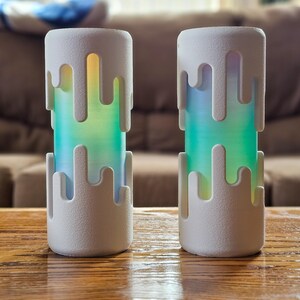 Può includere: Due lampade cilindriche bianche con un design a cera colante. Le lampade hanno un gradiente colorato di luce verde, blu e gialla. Sono posizionate su un tavolo di legno, con uno sfondo sfocato di un divano.