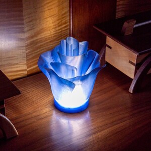 May include: A blue, flower-shaped decorative lamp with a glowing interior. The lamp is made of a translucent material, with the light source emanating from within. The lamp is placed on a wooden surface.