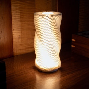 Puede incluir: Una lámpara cilíndrica blanca con un diseño de remolino y textura. La lámpara está encendida y proyecta un brillo cálido.