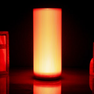 May include: A red cylindrical lamp with a white base and a ribbed glass shade. The lamp is turned on and emitting a warm red glow.
