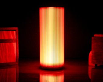 May include: A red cylindrical lamp with a white base and a ribbed glass shade. The lamp is turned on and emitting a warm red glow.
