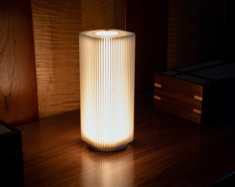 May include: A white cylindrical lamp with a pleated shade, illuminating a wooden surface. The lamp is turned on and casting a warm glow.