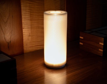 May include: A white cylindrical lamp with a geometric pattern on the shade. The lamp is turned on and illuminating the surrounding area.