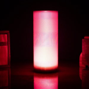 May include: A pink and white cylindrical lamp with a wavy pattern. The lamp is turned on and casting a warm glow.