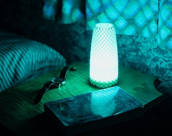 May include: A bedside table with a book, a watch, and a white lamp with a geometric pattern. The lamp is lit with a blue light, casting a blue glow on the table and the book.