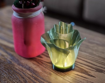 May include: A green 3D printed rose shaped candle holder with a warm white light inside. The candle holder is sitting on a wooden table next to a pink jar.