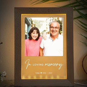 May include: A wooden photo frame with a warm glow, displaying a photo of a man and woman. The frame has the text "In loving memory" and "Father 1953 - 2022" inscribed on it.