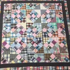 May include: A patchwork quilt made from squares of different plaid fabrics in shades of blue, pink, brown, and white. The quilt is surrounded by a black border.