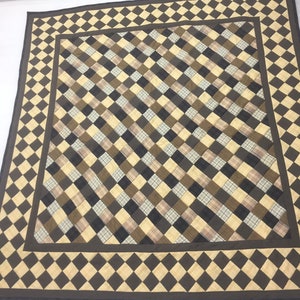 May include: A square quilted throw featuring a brown, beige, and black plaid pattern. The throw has a diamond-shaped border in brown and beige.