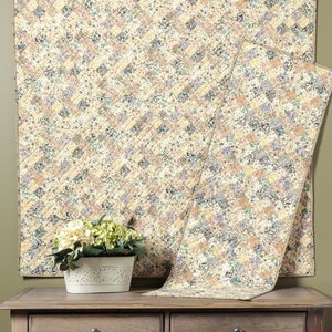 May include: Two quilted wall hangings with a floral pattern in shades of cream, beige, and blue. The smaller hanging is draped over a wooden table with a white planter and green plant.