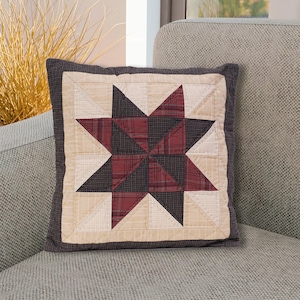 以下が含まれることがあります： 星形のデザインが施された、茶色、黒、クリーム色のキルトのクッション。クッションは灰色のソファの上に置かれています。