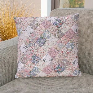 May include: A decorative cushion with a patchwork design featuring a variety of floral patterns in shades of pink, blue, and white. The cushion is sitting on a light grey sofa.