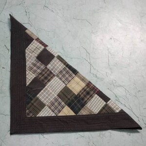 Puede incluir: Una colcha triangular de patchwork con un borde marrón. La colcha está hecha de varios tejidos de cuadros marrones, beige y verdes.