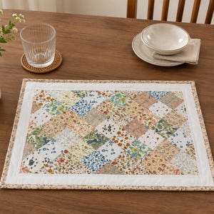 May include: A rectangular floral patchwork placemat with a white border. The placemat features a variety of floral patterns in shades of blue, green, pink, and yellow. A glass, coaster, bowl, plate, and napkin are on a wooden table.