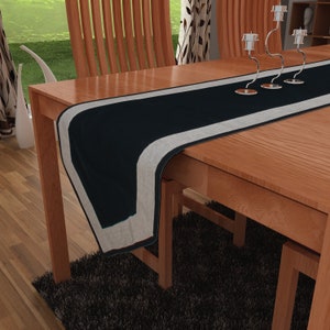 Op de afbeelding: Een zwart-witte tafelrunner met een grijze rand. De runner ligt op een houten tafel met twee stoelen.