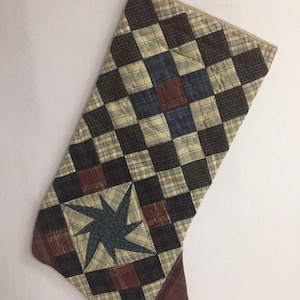 May include: A quilted Christmas stocking with a patchwork design featuring brown, blue, tan, and red plaid fabric. The stocking has a star-shaped design in the center.