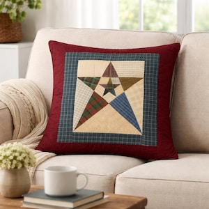 Op de afbeelding: Een vierkant decoratief kussen met een patchwork sterontwerp. Het heeft een bordeauxrode rand, een blauw en groen geruit vierkant en een centrale ster bestaande uit verschillende stofpatronen in beige, rood, groen en blauw. Het kussen ligt op een beige bank.
