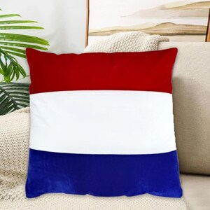Puede incluir: Un cojín cuadrado con un diseño tricolor en rojo, blanco y azul. El cojín está hecho de una tela suave y está dividido en tres franjas horizontales. El cojín está colocado sobre un sofá beige.