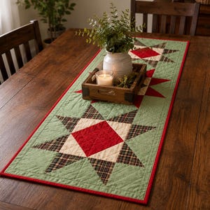 May include: A rectangular quilted table runner featuring a repeating star pattern in sage green, cream, and red, with dark plaid accents. The runner is finished with a solid red border and intricate diamond-shaped machine quilting throughout the fabric.