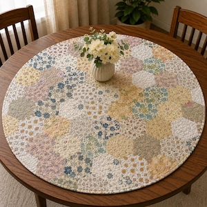 Op de afbeelding: Een rond gewatteerd tafelkleed met een patchwork van zeshoekige stoffen stukjes in diverse bloemmotieven met zachte tinten geel, blauw, roze en wit. In het midden staat een witte keramische vaas gevuld met witte bloemen.