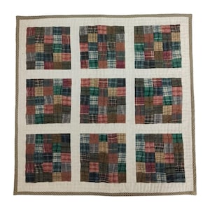 May include: A patchwork quilt featuring nine squares of plaid fabrics in red, green, brown, and blue. The squares are separated by white fabric, with a patterned trim border. The quilt is a cozy home decor item.