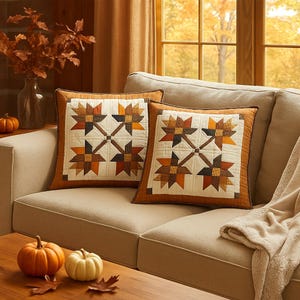 May include: Two square cushions with a patchwork star design in shades of brown, orange, and cream. The cushions are on a beige sofa, with pumpkins and autumn foliage nearby. The cushions have a brown border.