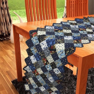 May include: A quilted table runner with a patchwork design in shades of blue, gray, and white. The runner is placed on a wooden table, adding a decorative touch to the dining area. The runner's pattern includes floral and geometric elements.
