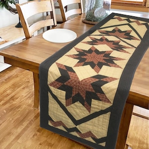 May include: A quilted table runner with a black, beige, and red star pattern. The runner has a black border and is on a wooden table.
