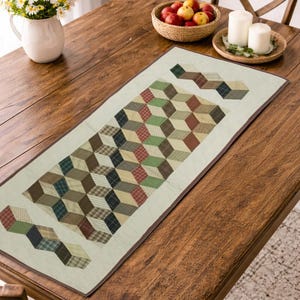 May include: A rectangular table runner with a geometric cube pattern in shades of brown, green, and red. The runner is placed on a wooden table, with a vase of flowers and a bowl of fruit nearby. The runner is approximately 12 inches wide.
