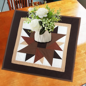 May include: A brown and beige quilted table runner with a star design. The runner has a dark brown border and is decorated with a white vase of white flowers.