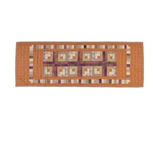 May include: A rectangular quilted table runner in shades of orange, brown, and purple. The center features a pattern of squares within squares, surrounded by a border of alternating colors and patterns. The runner is made of fabric.