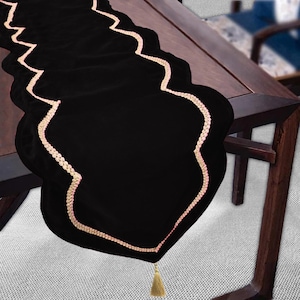 Puede incluir: Camino de mesa de terciopelo negro con borde festoneado, ribeteado con un borde decorativo color crema. Una borla dorada cuelga de la parte inferior. El camino de mesa está sobre una mesa de madera oscura.