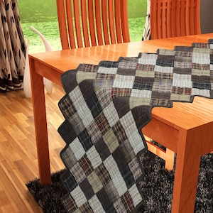 May include: A brown, black, and white patchwork table runner with a quilted design. The runner is on a wooden table with two chairs in the background.