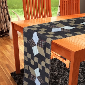 Puede incluir: Camino de mesa rectangular con diseño de patchwork. El camino de mesa presenta un patrón de formas geométricas negras, beige y blancas, con secciones de tela a cuadros y a rayas. Se muestra sobre una mesa de madera.
