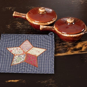 Könnte beinhalten: Zwei braune Keramik-Aufläufe mit Deckeln, einer mit Griff, auf einem blauen und weißen gesteppten Topflappen mit einem sternförmigen Patchwork-Design.