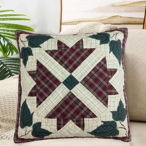 Puede incluir: Cojín decorativo cuadrado con un estampado de cuadros en burdeos, crema y verde azulado. El cojín presenta un diseño central de estrella y está bordeado con hojas de color verde oscuro. El cojín es probablemente para la decoración del hogar.