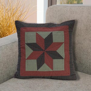 May include: Square cushion with a star quilt design. The cushion has a dark grey border, a red square, and a star pattern in red, green, and dark grey. The cushion is placed on a light grey sofa.