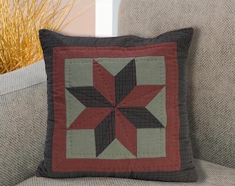 Puede incluir: Cojín decorativo cuadrado con diseño de edredón de estrella. El cojín presenta un borde gris oscuro, un cuadrado rojo y un patrón de estrella en rojo, verde y gris oscuro. El cojín está colocado en un sofá gris claro.